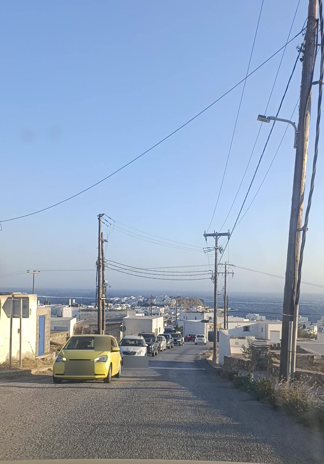 Στενός δρόμος με σταθμευμένα και σταματημένα οχήματα, η Χώρα και η θάλασσα στο βάθος