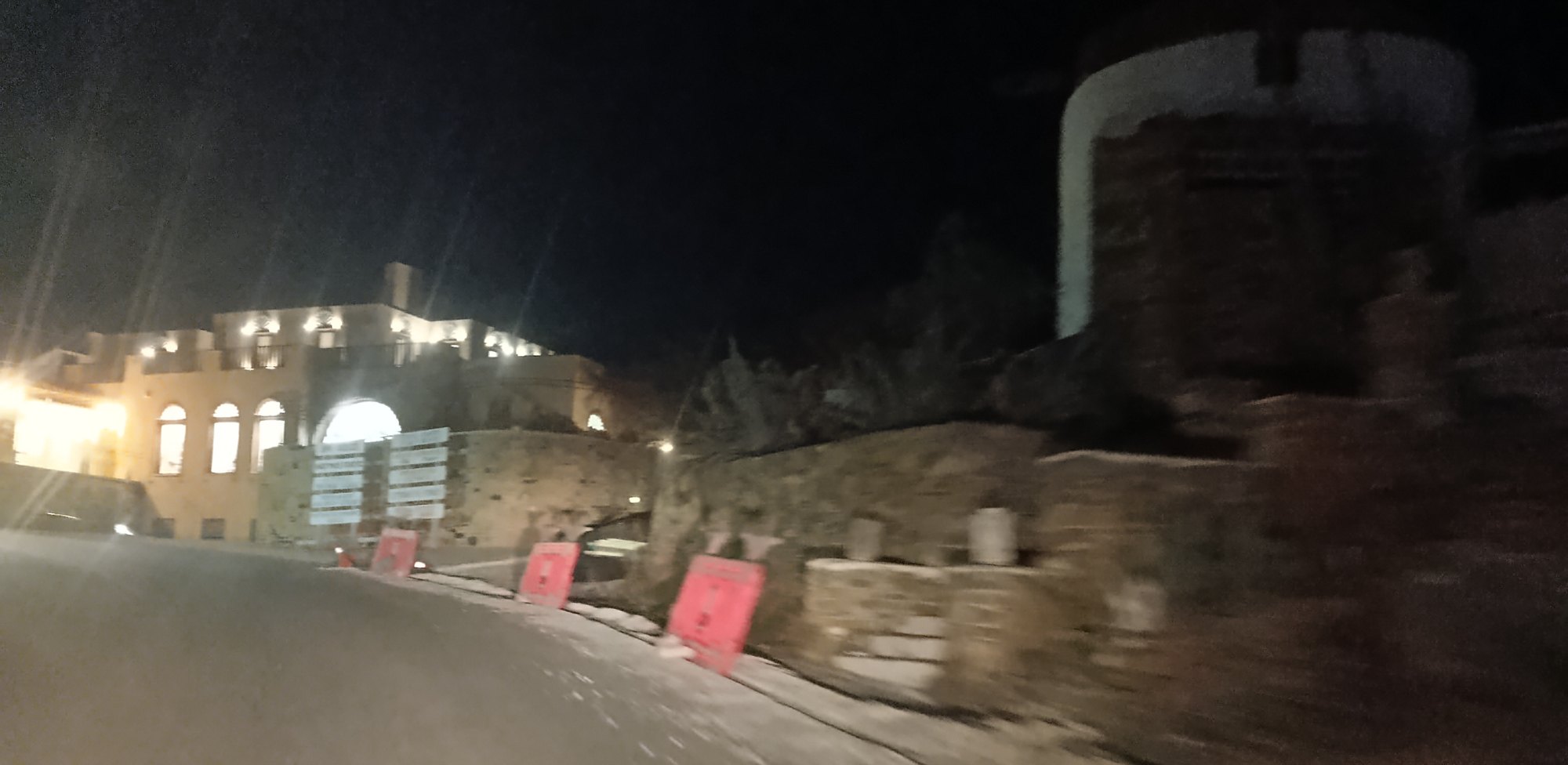 Κόκκινα κιγκλιδώματα σε σειρά μπροστά από ξερολιθιά, με τον ανεμόμυλο και φωτισμένο κτίριο στο βάθος, τη νύχτα