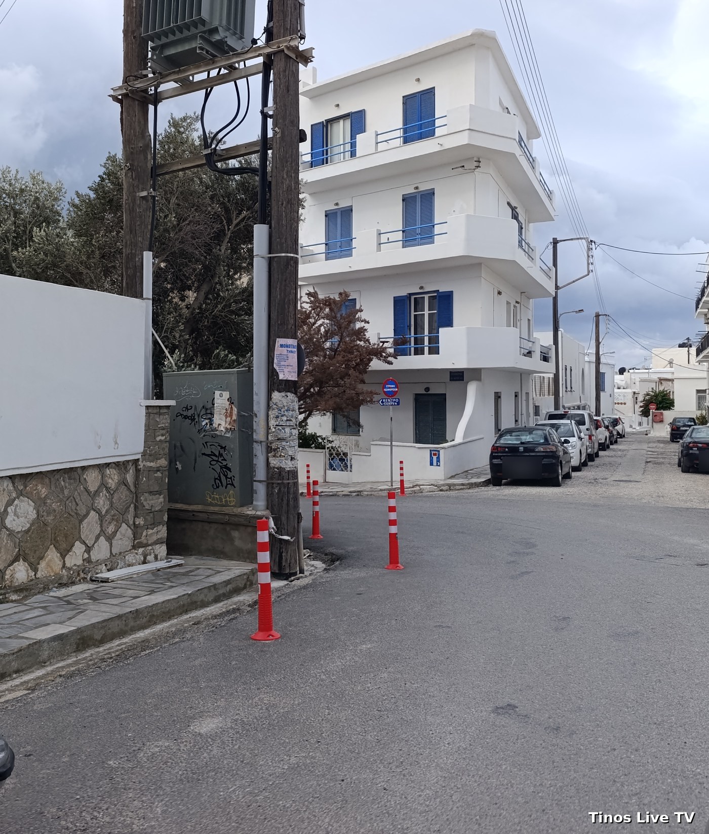 Η γωνία Αγίου Ιωάννου – 25ης Μαρτίου στη Χώρα της Τήνου σήμερα, με τέσσερα κόκκινα-λευκά κολωνάκια γύρω από τη γωνία δίπλα στην κολόνα του μετασχηματιστή