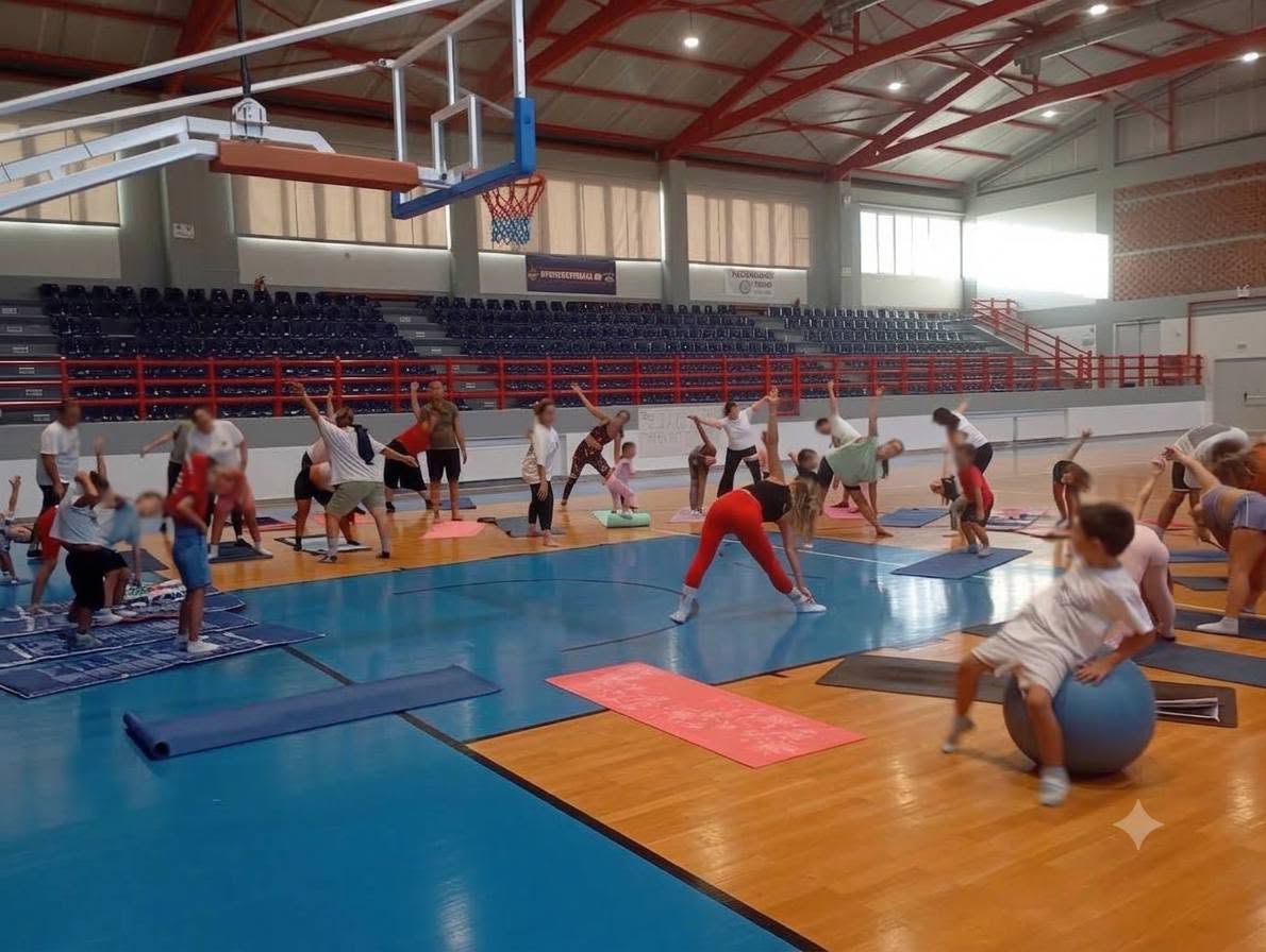 Γονείς και παιδιά γυμνάζονται όρθιοι με τα χέρια ψηλά στο κλειστό γυμναστήριο Τήνου