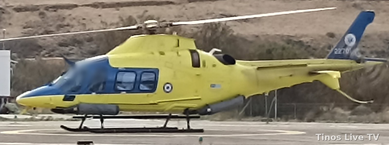 Κίτρινο ελικόπτερο αεροδιακομιδών AW109 Trekker με το εθνικό σήμα στο ελικοδρόμιο της Τήνου