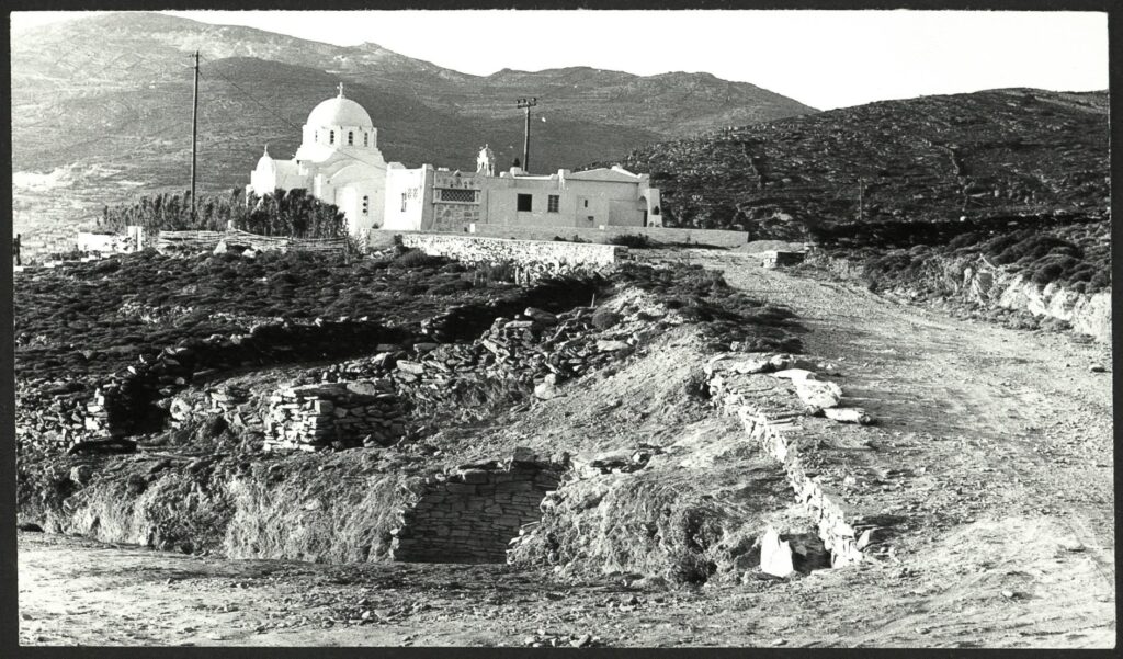 Ασπρόμαυρη φωτογραφία της ανασκαφής του 1979: ο θολωτός τάφος δίπλα στον χωματόδρομο και στο βάθος ο ναός της Αγίας Θέκλας