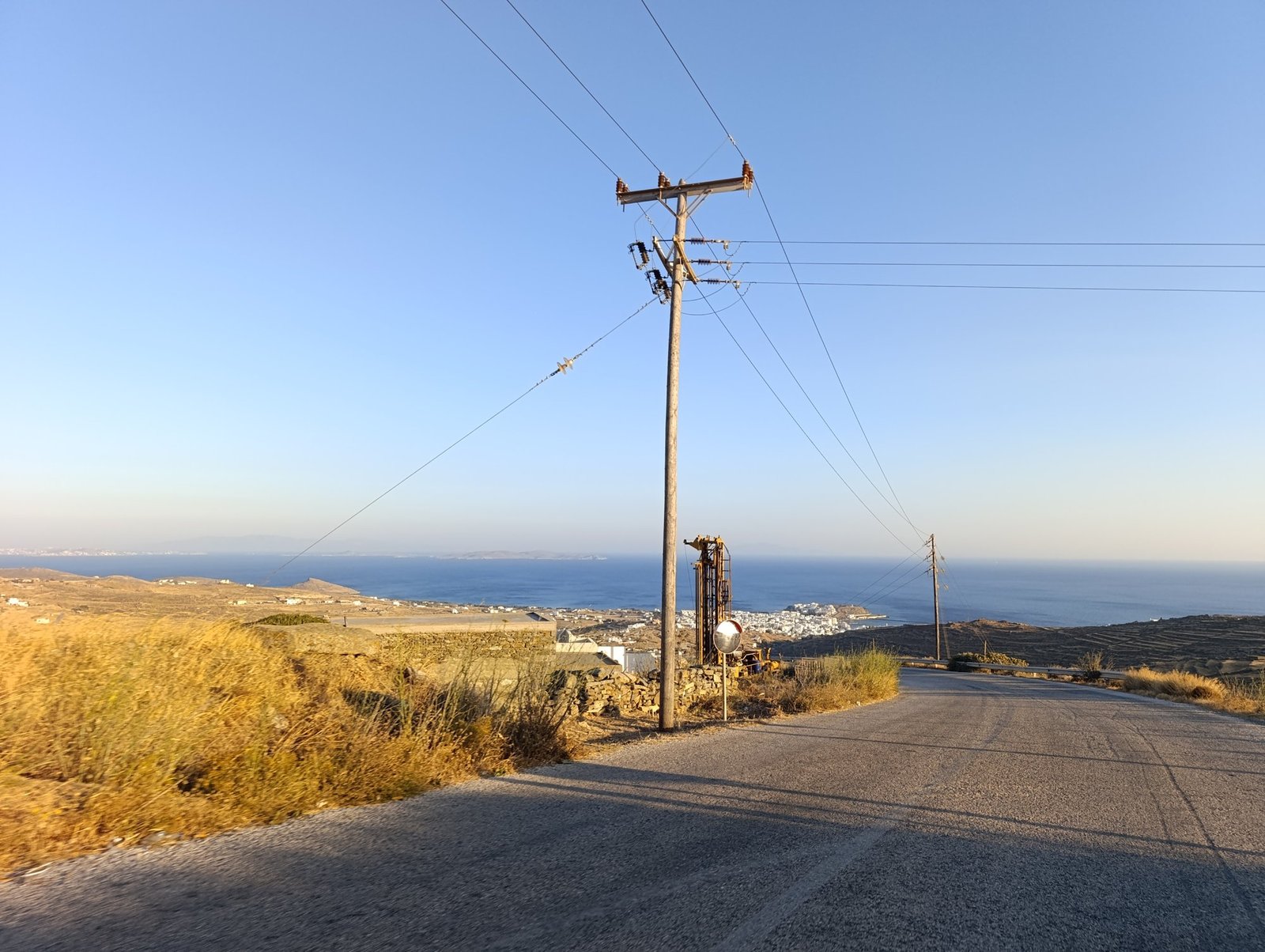 Κολόνα του δικτύου διανομής ηλεκτρικής ενέργειας σε δρόμο της Τήνου, με θέα προς τη Χώρα και τη θάλασσα