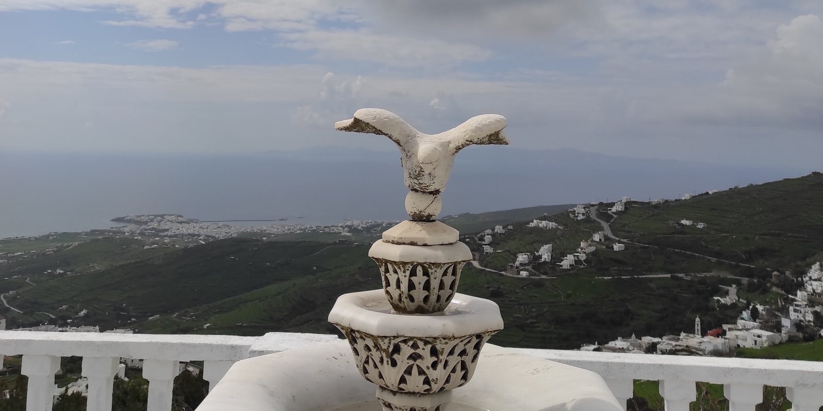 Το μαρμάρινο γλυπτό με το πουλί απέναντι από τον ναό της Κοιμήσεως στα Δύο Χωριά Τήνου, με θέα ως τη Χώρα