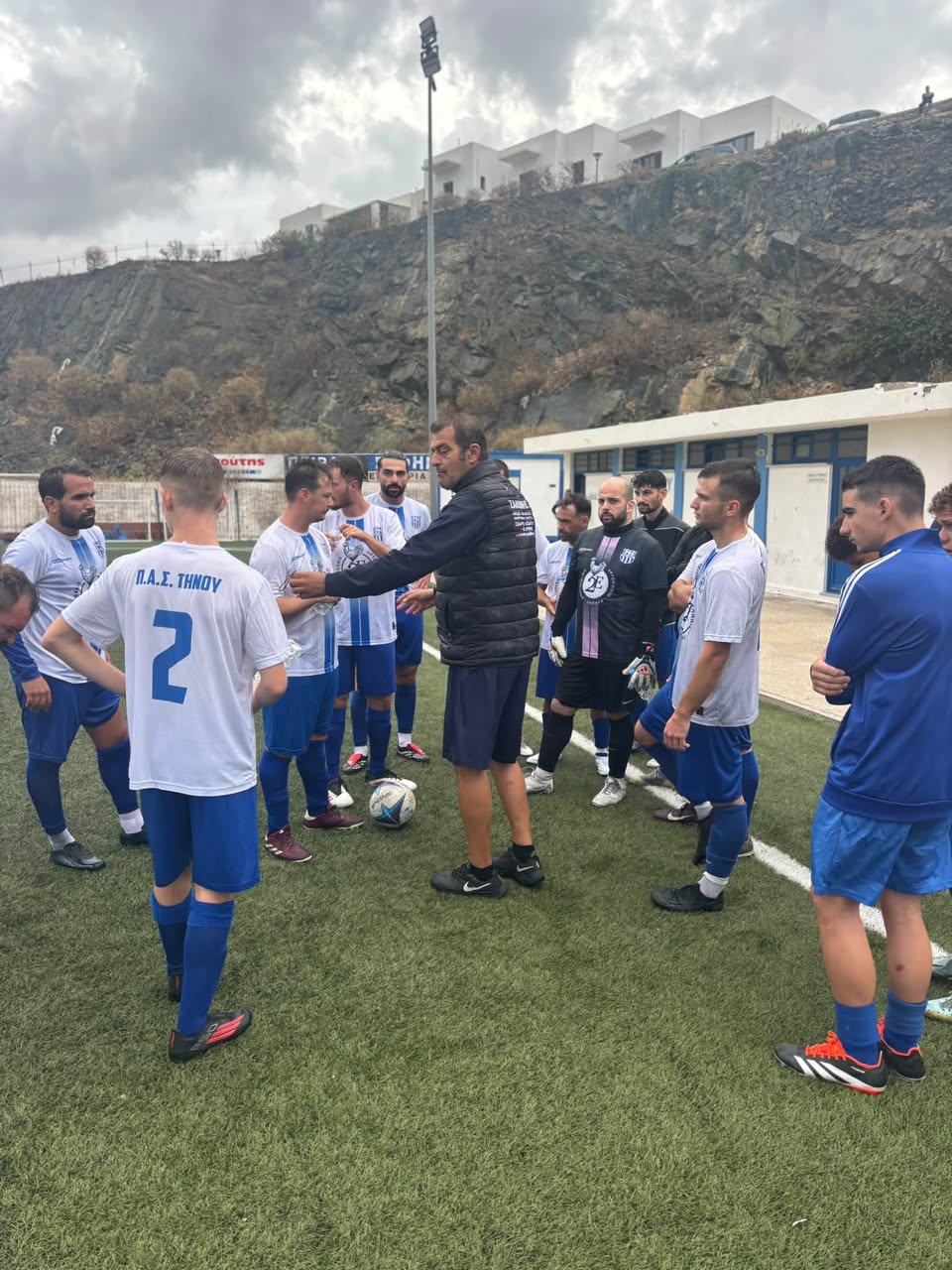 Οι ποδοσφαιριστές του Π.Α.Σ. Τήνου με λευκές φανέλες και μπλε σορτς, στον πάγκο του γηπέδου της Τήνου