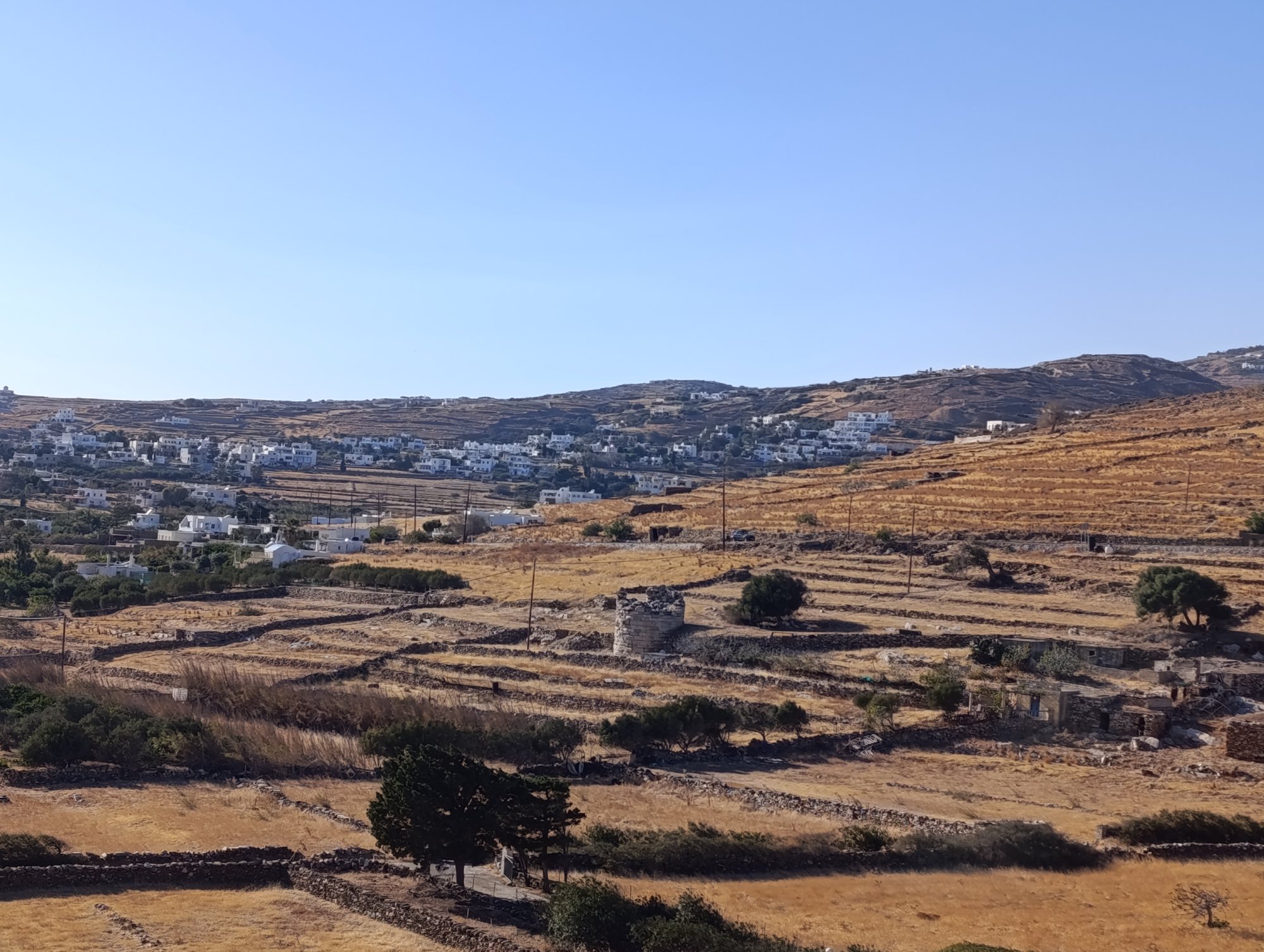 Ο αρχαίος κυλινδρικός πύργος του Σμόβολου στην Τήνο, ανάμεσα σε αναβαθμίδες με ξερολιθιές και ξερό χορτάρι, με λευκά σπίτια στην πλαγιά πίσω