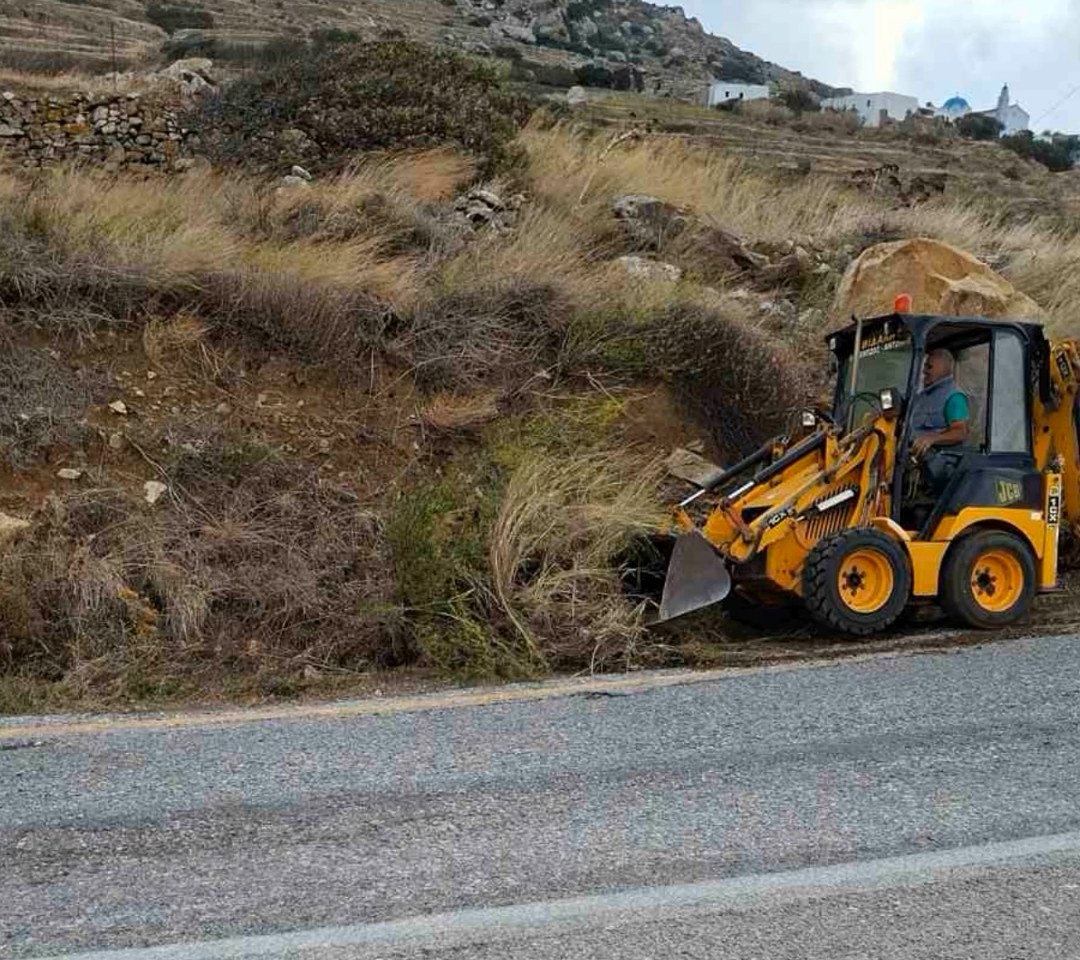 Μηχάνημα έργων καθαρίζει χόρτα στο πρανές δίπλα στον δρόμο, κάτω από βραχώδη λόφο