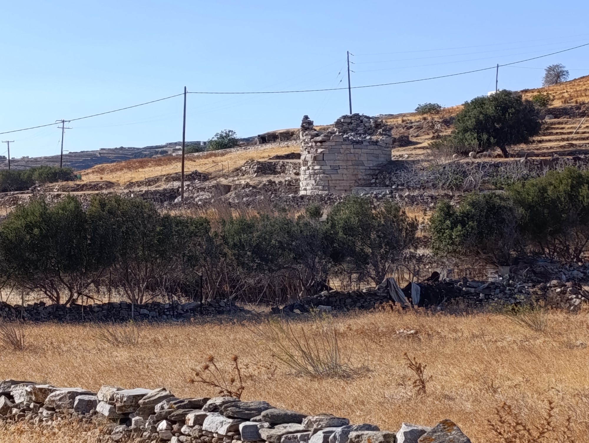 Ο πύργος του Σμόβολου από πιο κοντά: σειρές από ορθογώνιους μαρμάρινους δόμους στο κάτω μέρος, πρόχειρη ξερολιθιά στην κορυφή, κολόνες ρεύματος και πρόβατα στη σκιά δέντρου