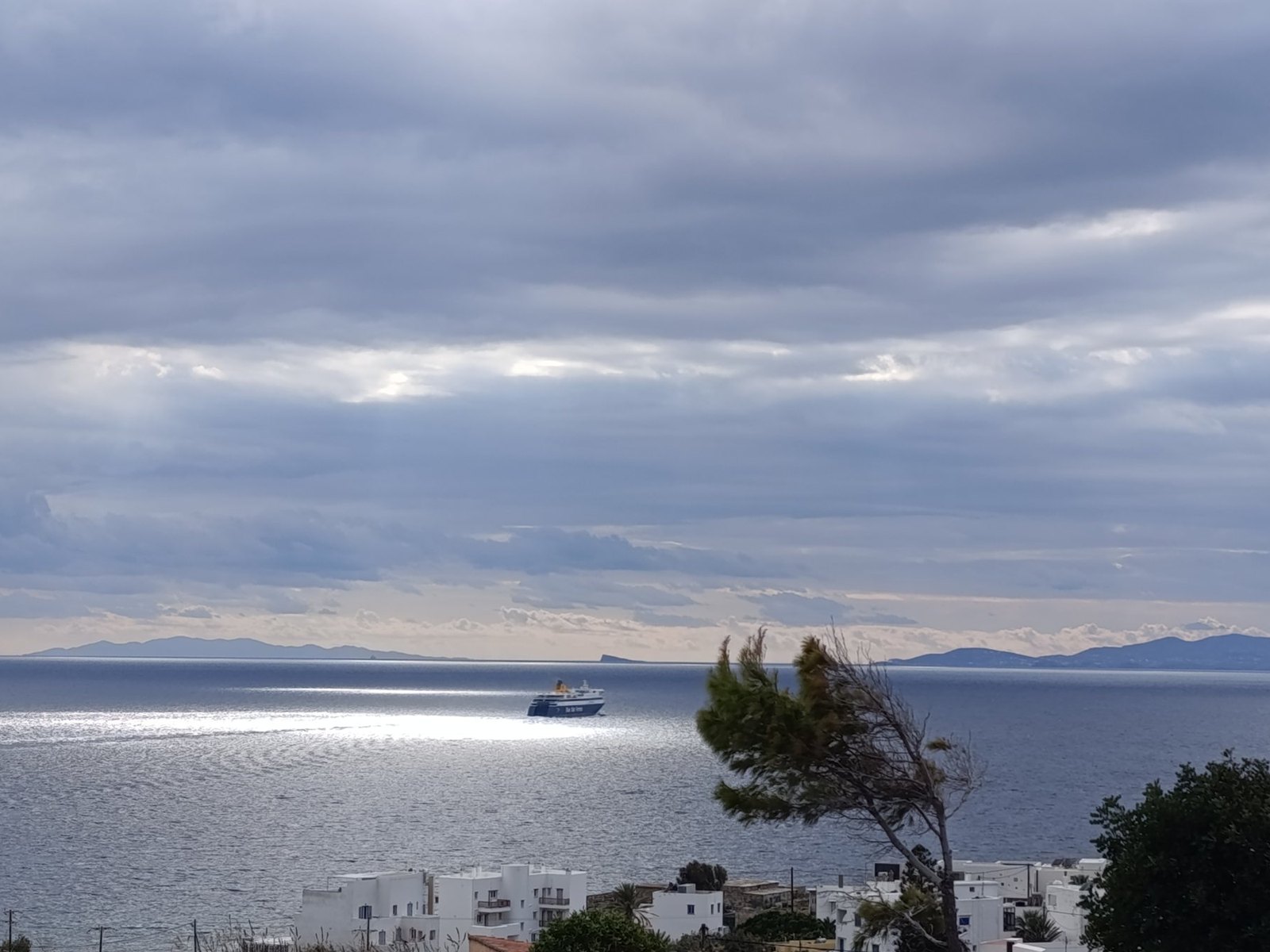 Το πλοίο Μπλου Σταρ Πάρος ανοιχτά της Τήνου, με γκρίζα σύννεφα και ηλιαχτίδες στη θάλασσα