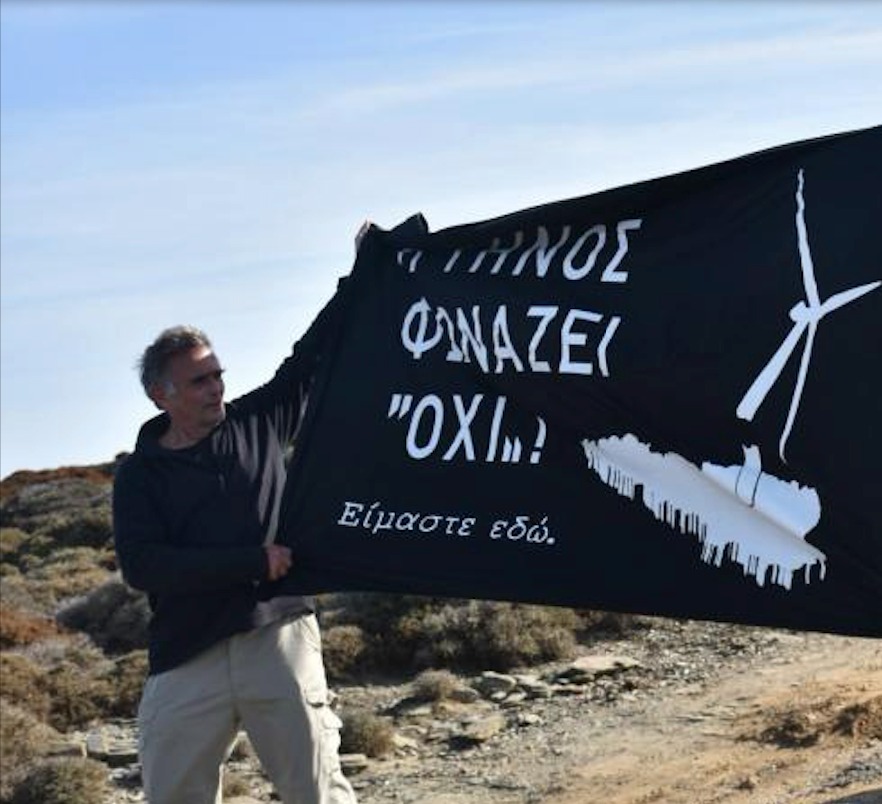 Ο Δημήτρης Παυλάκης κρατά πανό με το σύνθημα «Η Τήνος φωνάζει ΟΧΙ! Είμαστε εδώ» και σχέδιο ανεμογεννήτριας