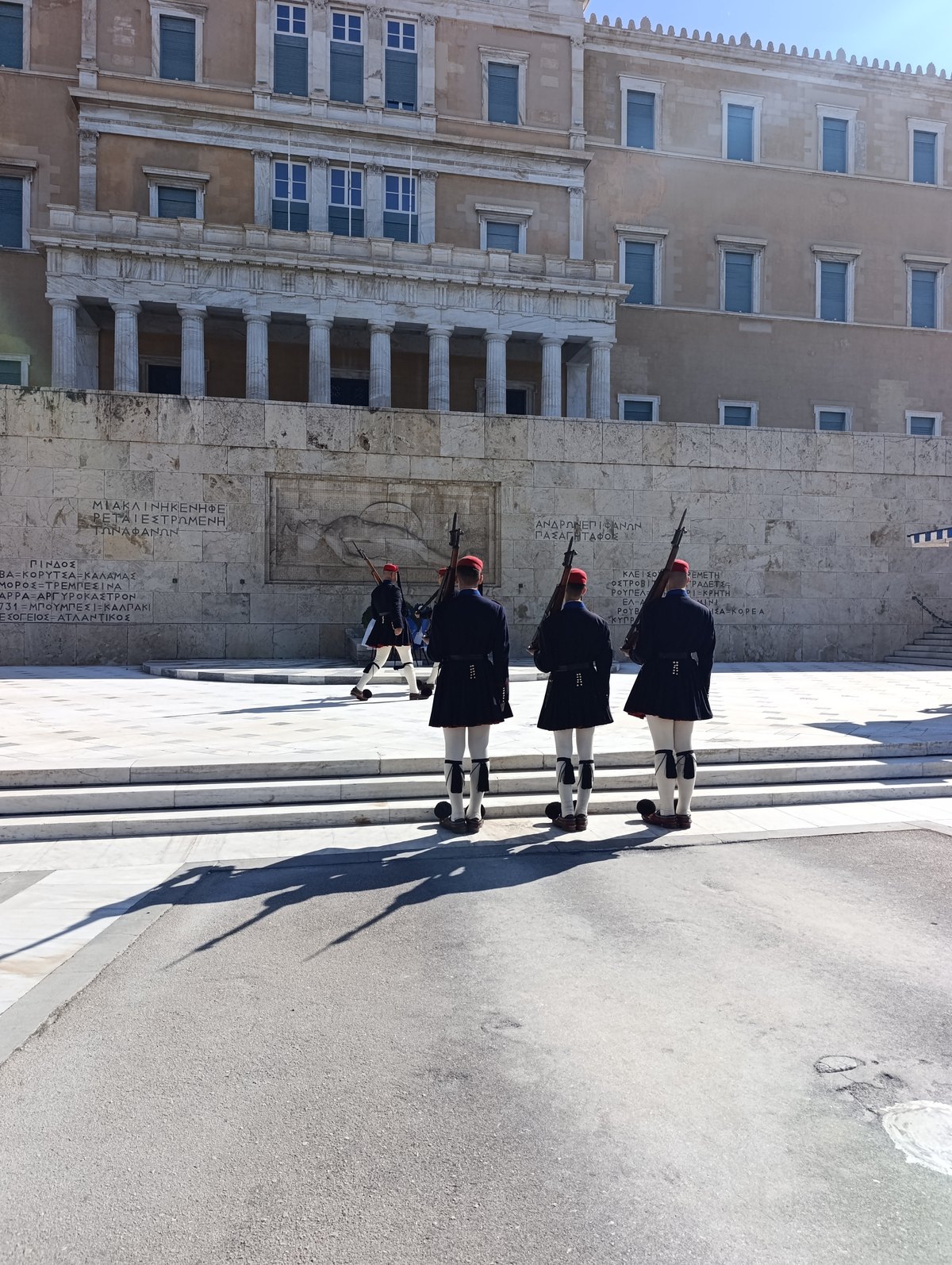 Τρεις Εύζωνες από πίσω μπροστά στο ανάγλυφο του Άγνωστου Στρατιώτη, με τα ονόματα των μαχών χαραγμένα στον τοίχο