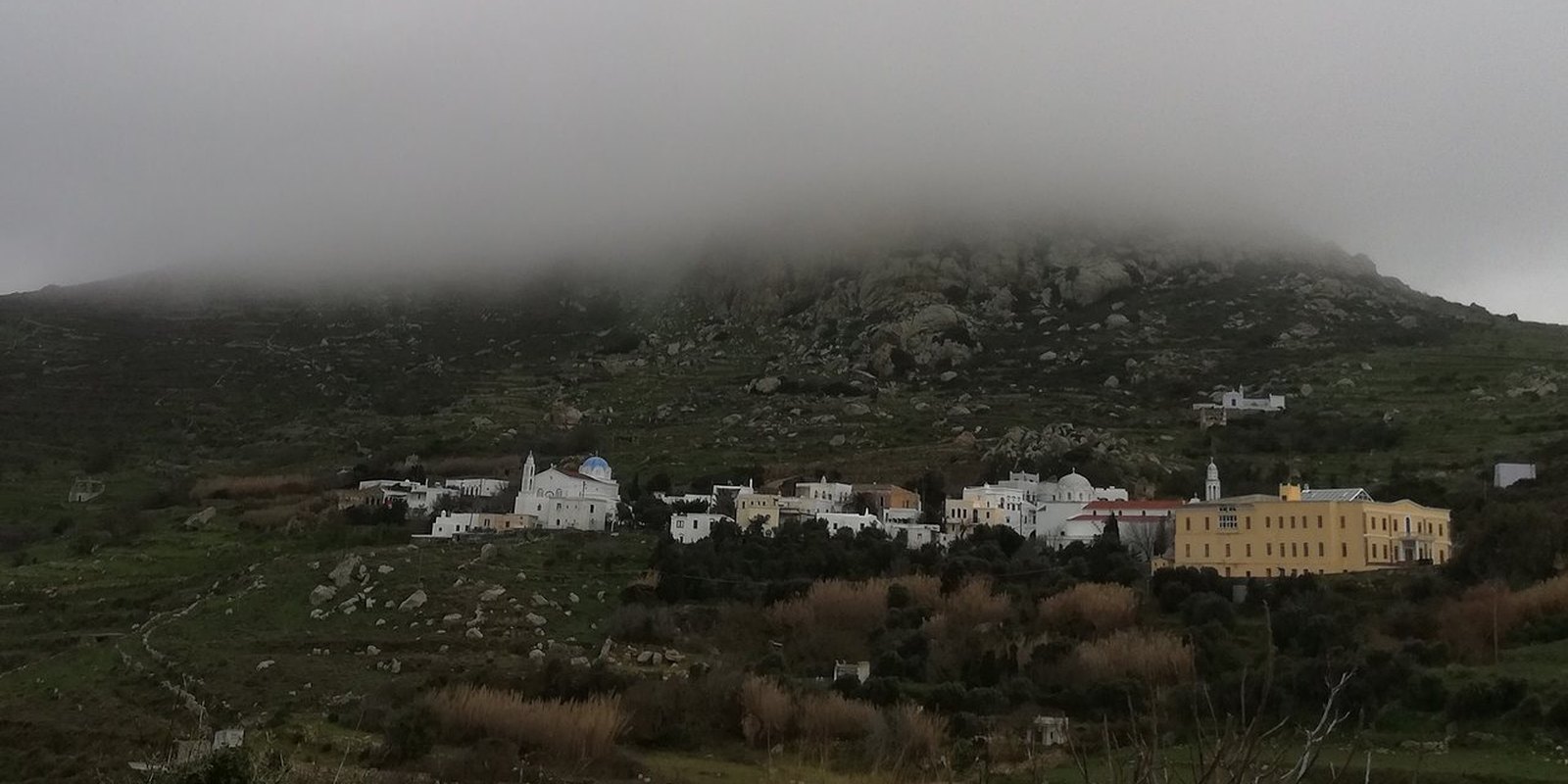 Η κορυφή του Εξωμβούργου χαμένη σε πυκνή ομίχλη, με το χωριό και τα κτίρια στην πράσινη πλαγιά