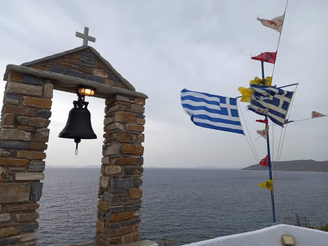 Καμπαναριό από πέτρα και ελληνικές σημαίες πάνω από τη θάλασσα της Τήνου