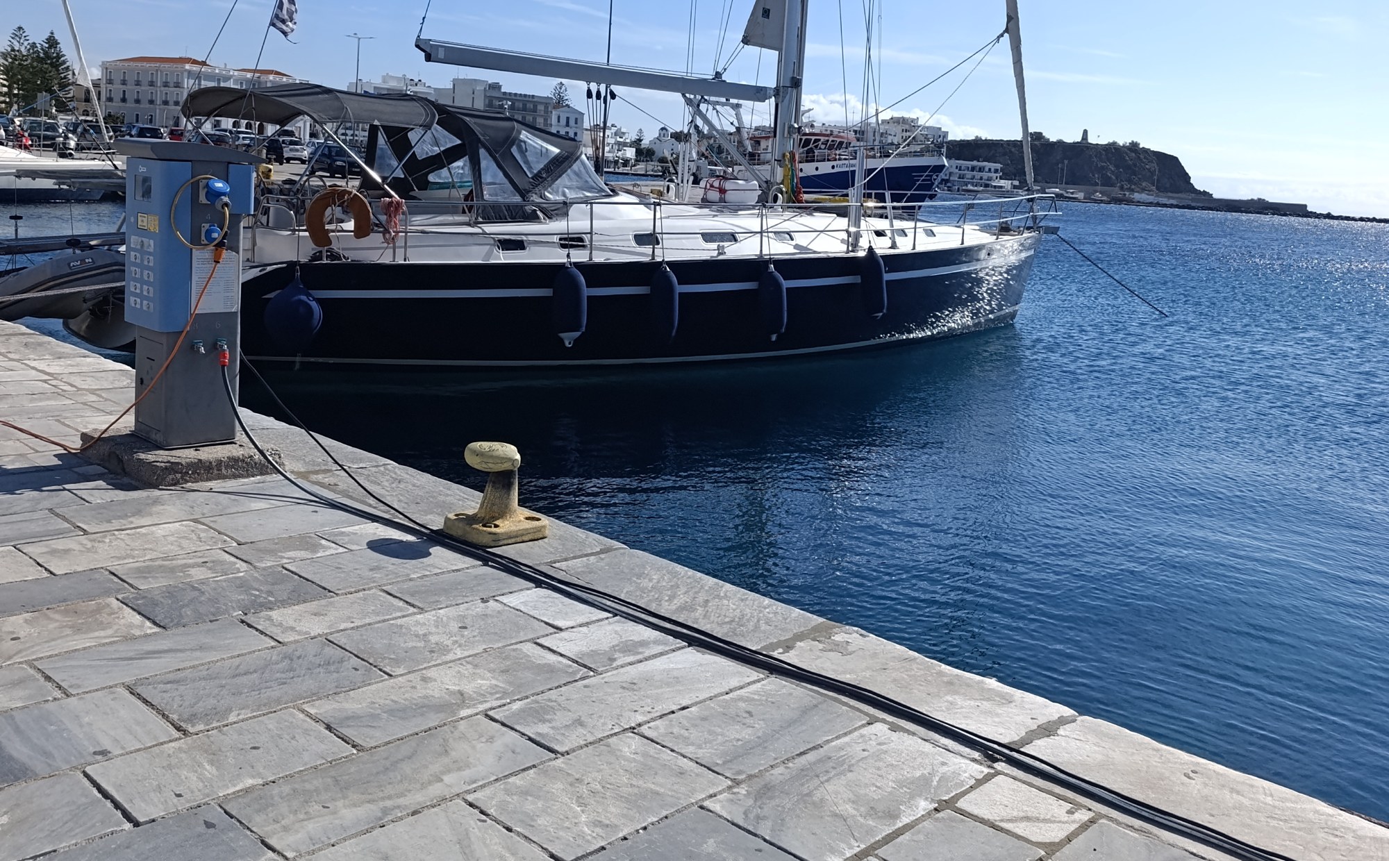 Ιστιοπλοϊκό με σκούρο μπλε κύτος δεμένο στον μαρμάρινο μόλο του λιμανιού της Τήνου, με ήρεμη θάλασσα