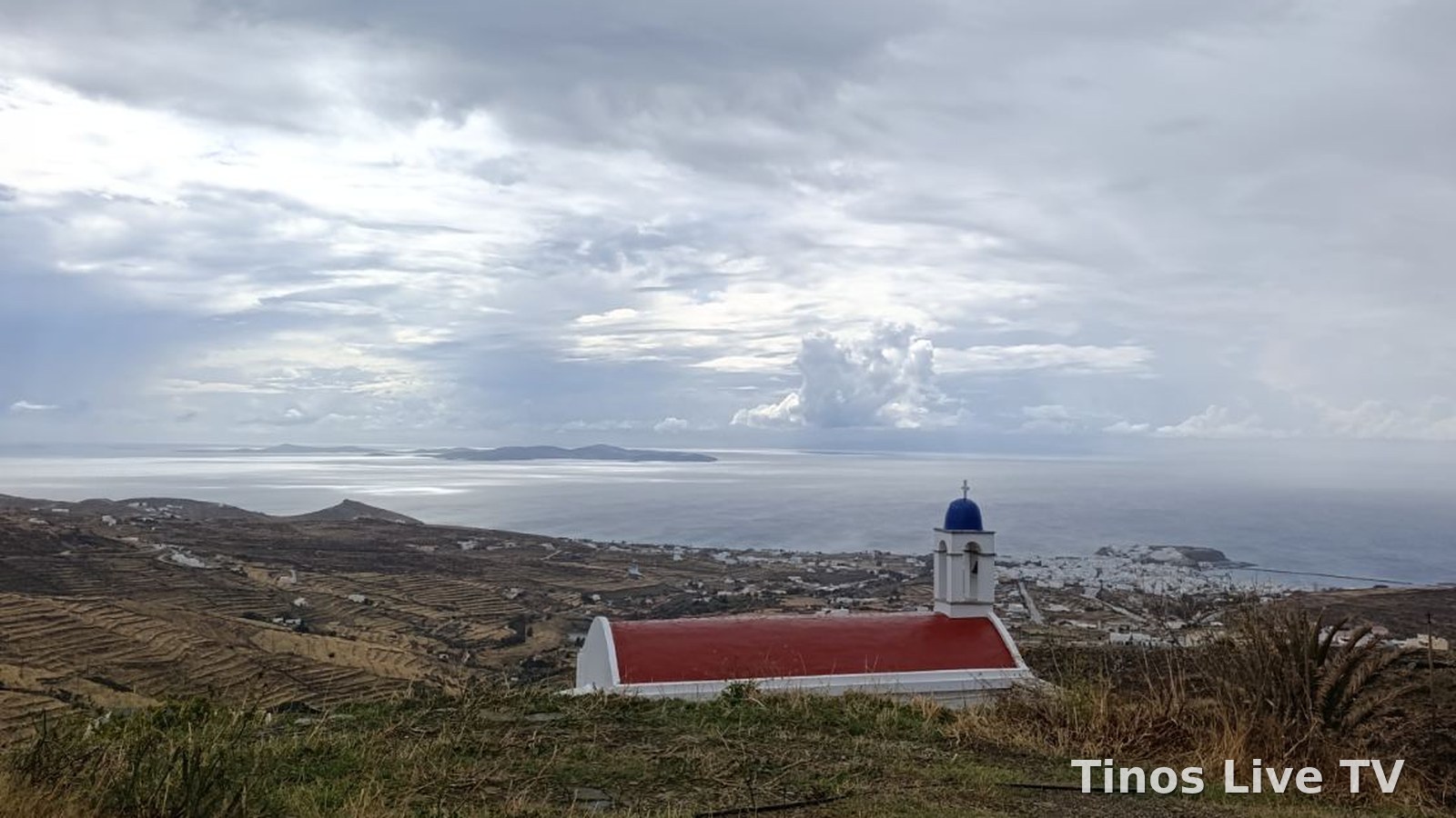 Εκκλησάκι με κόκκινη στέγη και μπλε τρούλο πάνω από τη Χώρα της Τήνου, κάτω από βαριά σύννεφα