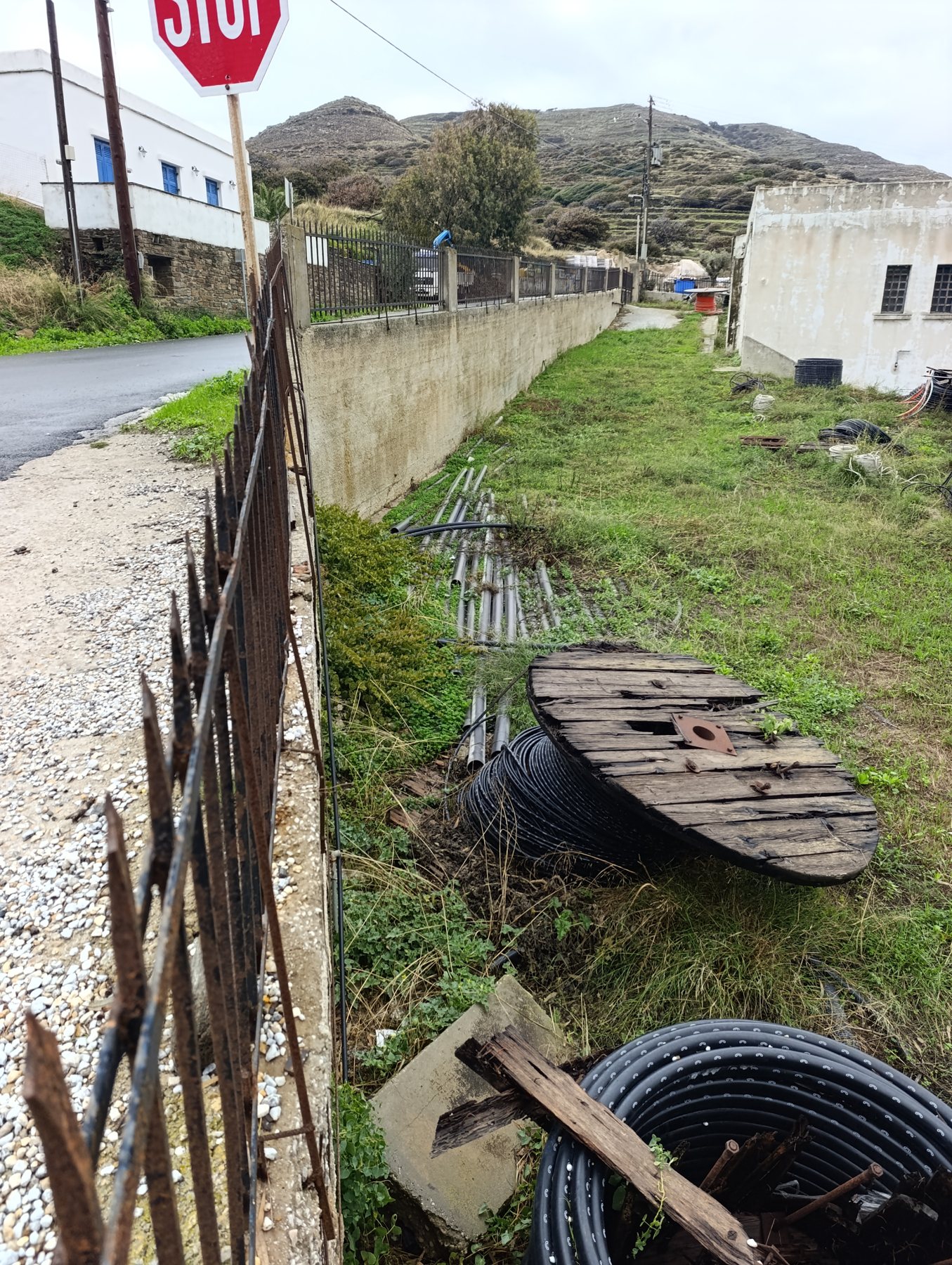 Σωλήνες, καλώδια και ξύλινες μπομπίνες δίπλα στον δρόμο, πίσω από σκουριασμένο κάγκελο, στην Κώμη Τήνου