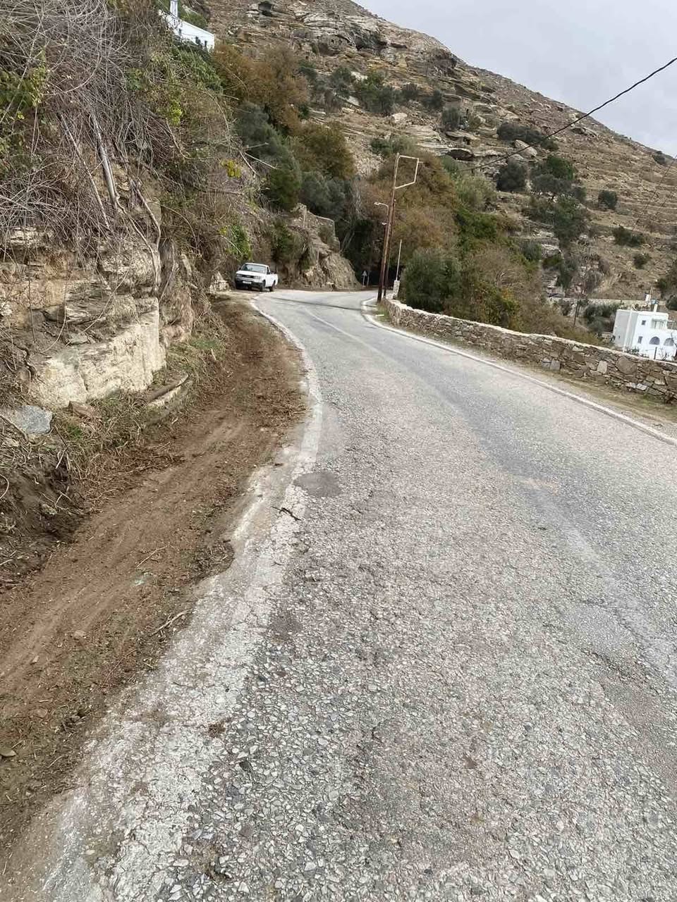Στροφή του επαρχιακού δρόμου στην Καρδιανή Τήνου με καθαρισμένο έρεισμα και χαντάκι, με τα σπίτια του χωριού στο βάθος