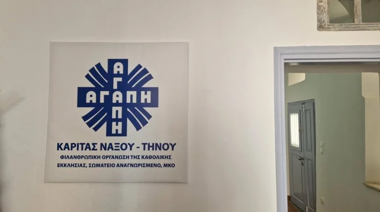 Η πινακίδα της Κάριτας Νάξου – Τήνου με το σήμα «ΑΓΑΠΗ» στον τοίχο, δίπλα σε ανοιχτή πόρτα