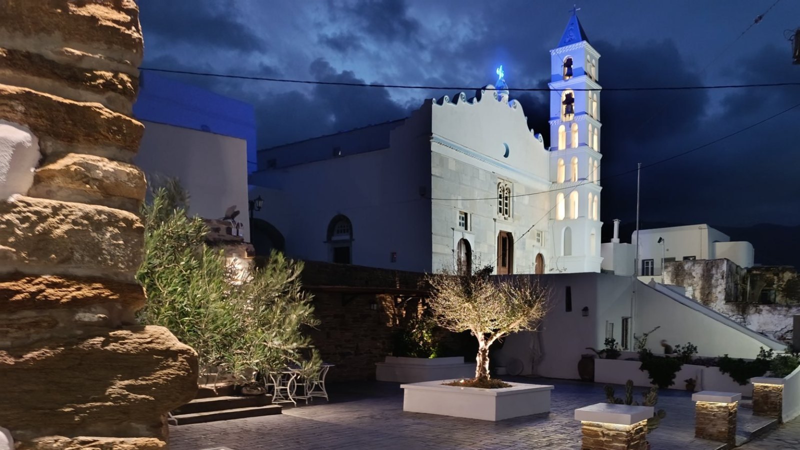 Η φωτισμένη πλατεία της Κάτω Κώμης το βράδυ, με την ελιά και το καμπαναριό του ναού του Αγιότατου Ονόματος της Παναγίας