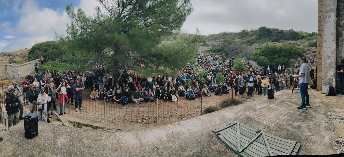 Πλήθος επισκεπτών κάθεται και ακούει την ξενάγηση ανάμεσα σε πεύκα και ερείπια στη Μακρόνησο