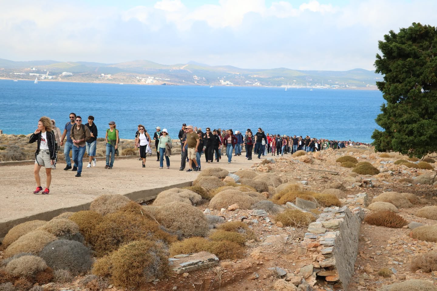 Σειρά επισκεπτών περπατά σε μονοπάτι της Μακρονήσου με τη θάλασσα και τις ακτές της Αττικής στο βάθος