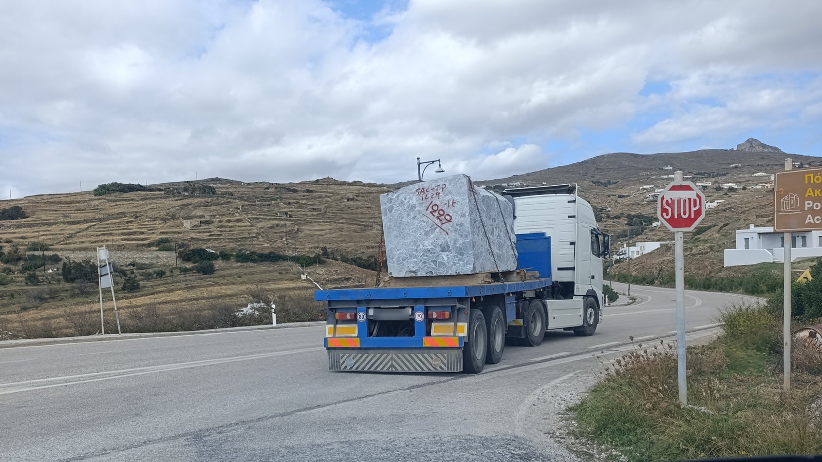 Νταλίκα με έναν τεράστιο όγκο μαρμάρου στρίβει σε δρόμο της Τήνου, με το Εξώμβουργο στο βάθος
