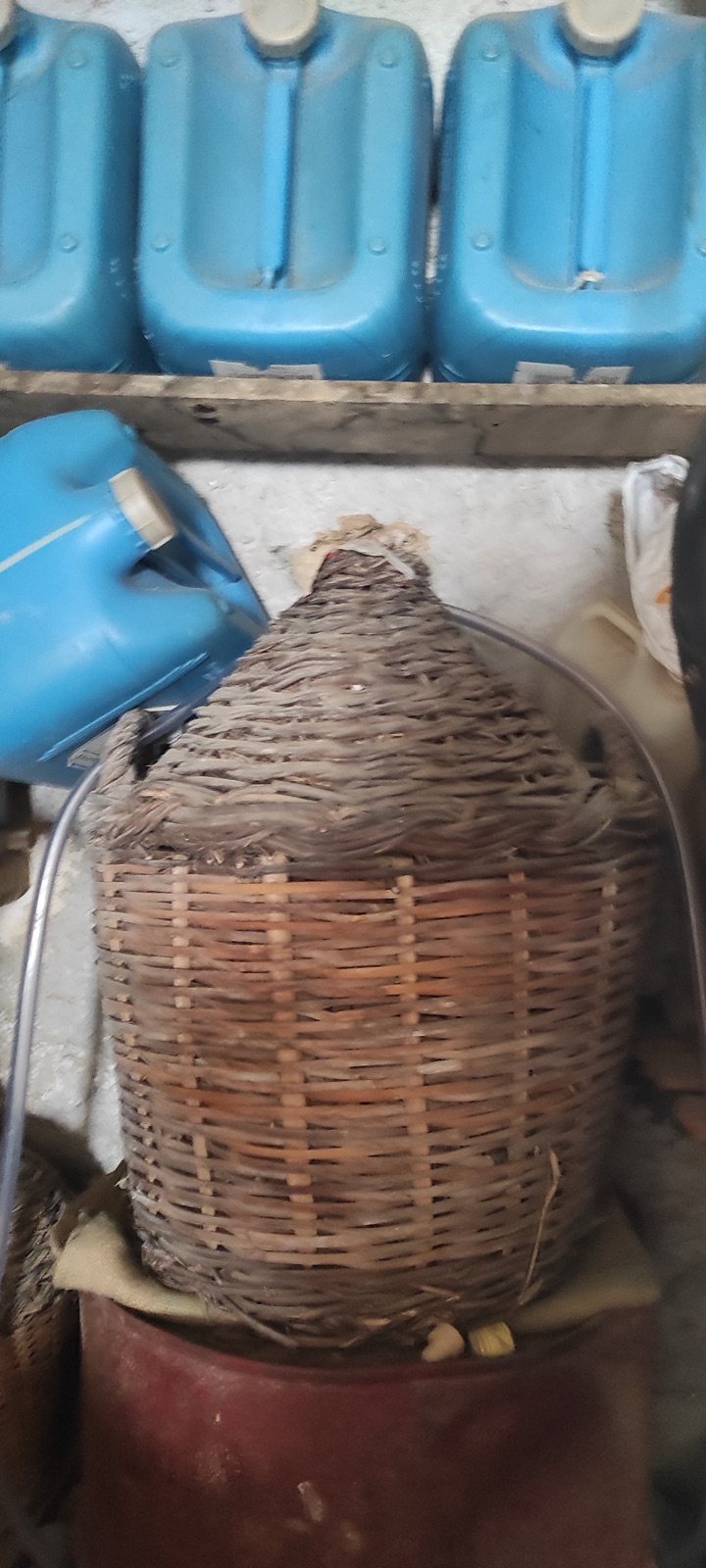 Μεγάλη ψάθινη νταμιτζάνα σε κατώι, με μπλε μπιτόνια στο ράφι από πάνω