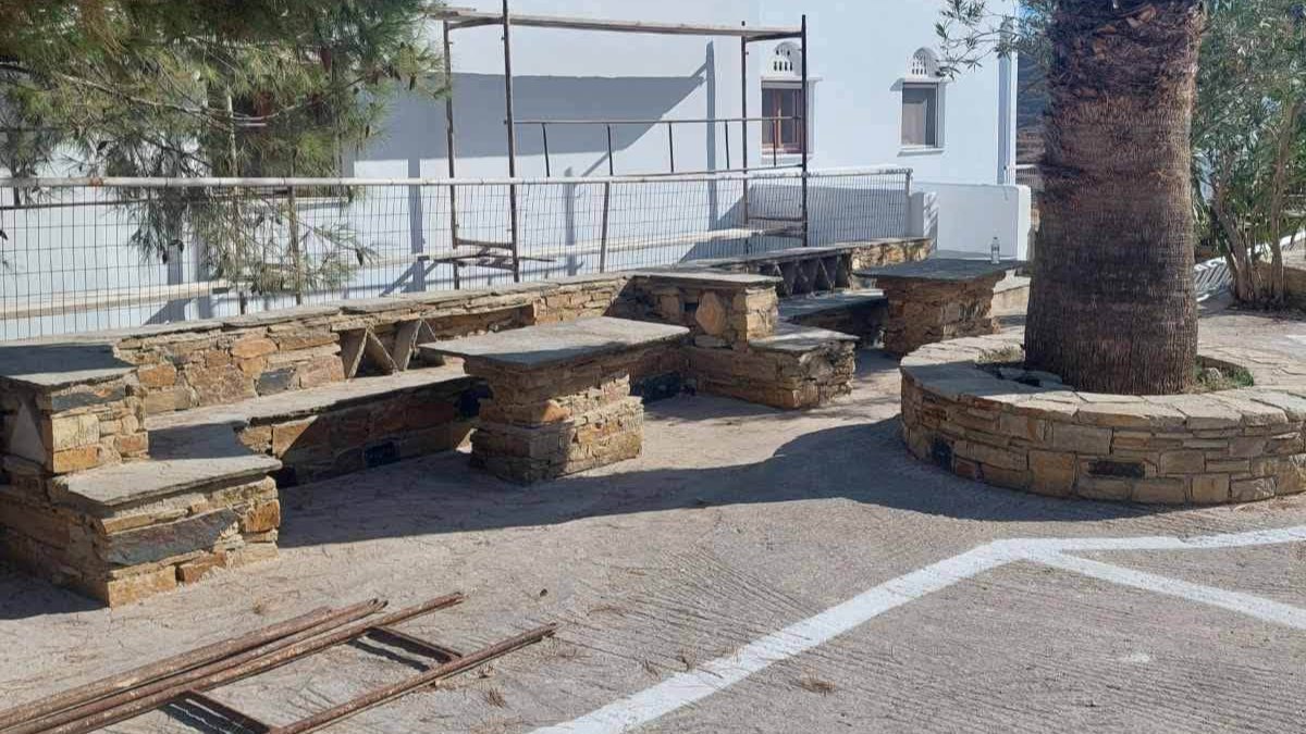 Η αυλή με τον φοίνικα και τα πέτρινα τραπέζια μπροστά στους ξενώνες της Παναγίας της Βουρνιώτισσας στην Τήνο