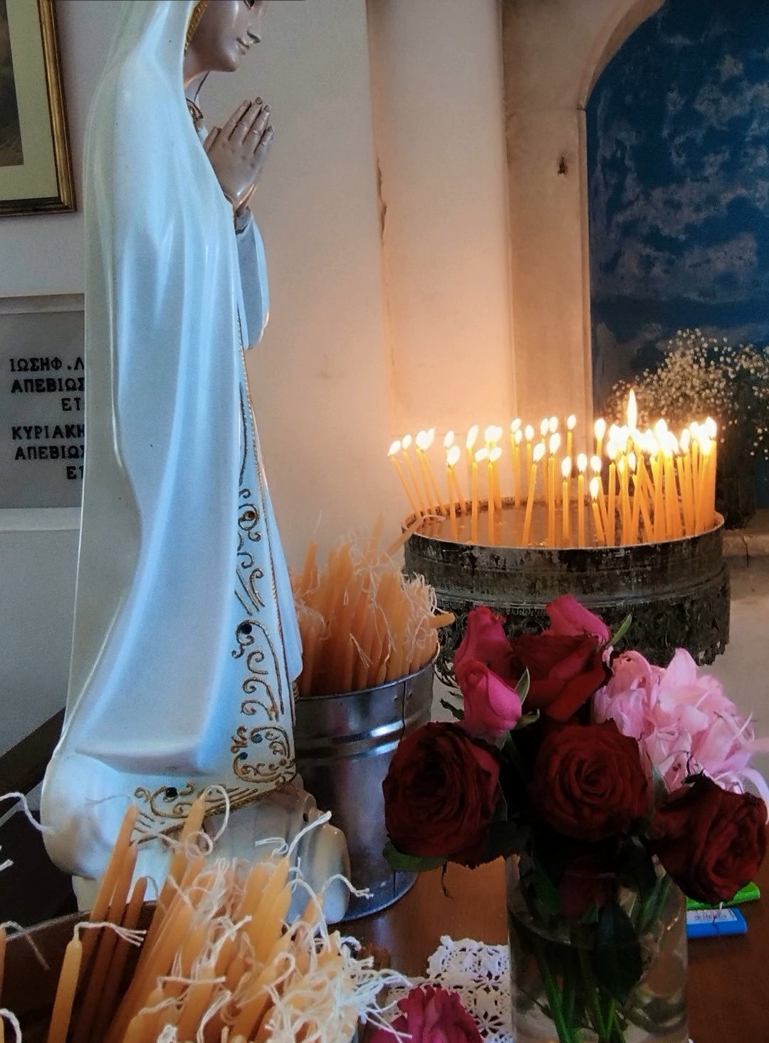 Άγαλμα της Παναγίας με αναμμένα κεριά και τριαντάφυλλα