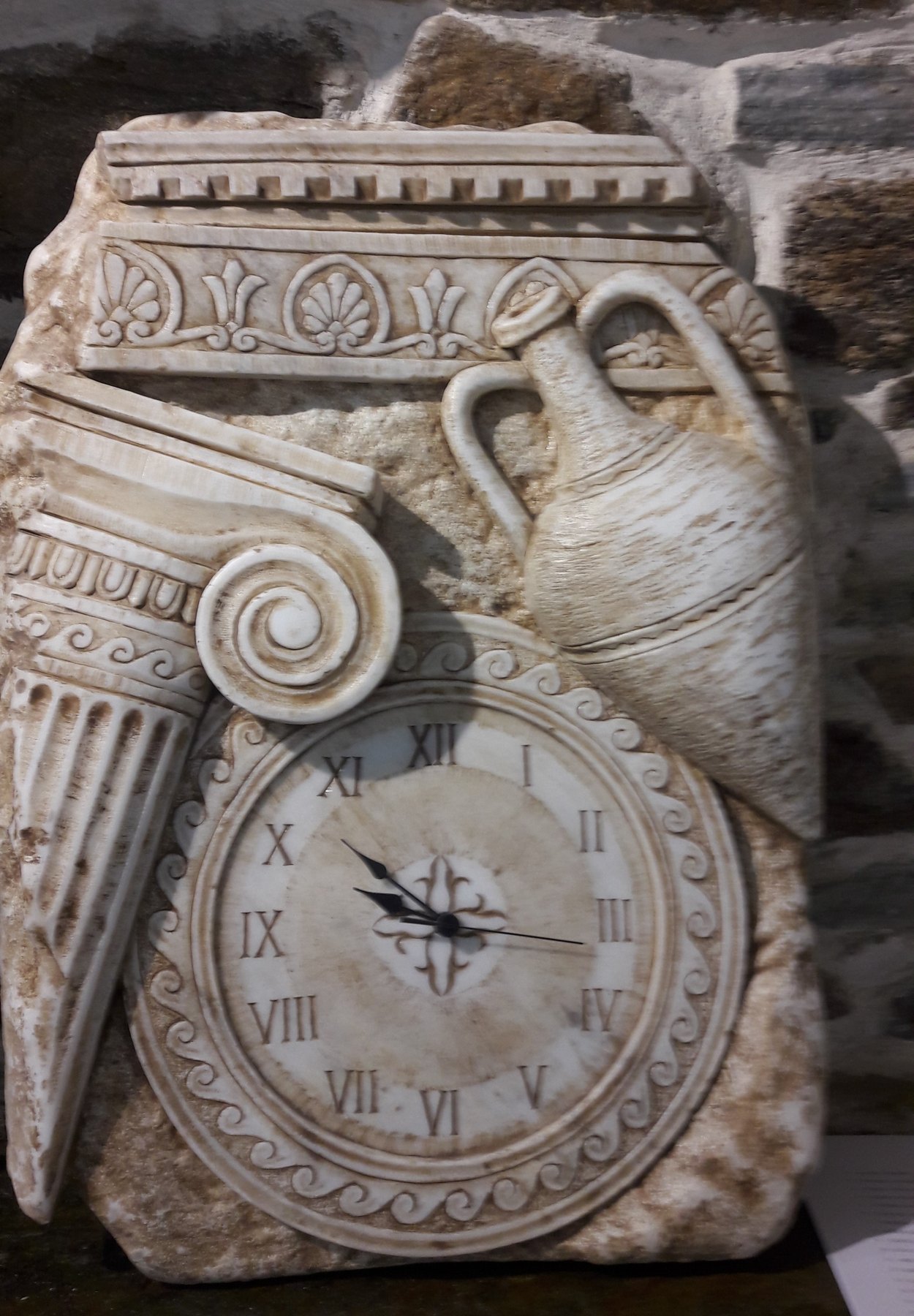 Ρολόι σε ανάγλυφο με ιωνικό κιονόκρανο και αμφορέα, μπροστά σε πέτρινο τοίχο στην Τήνο