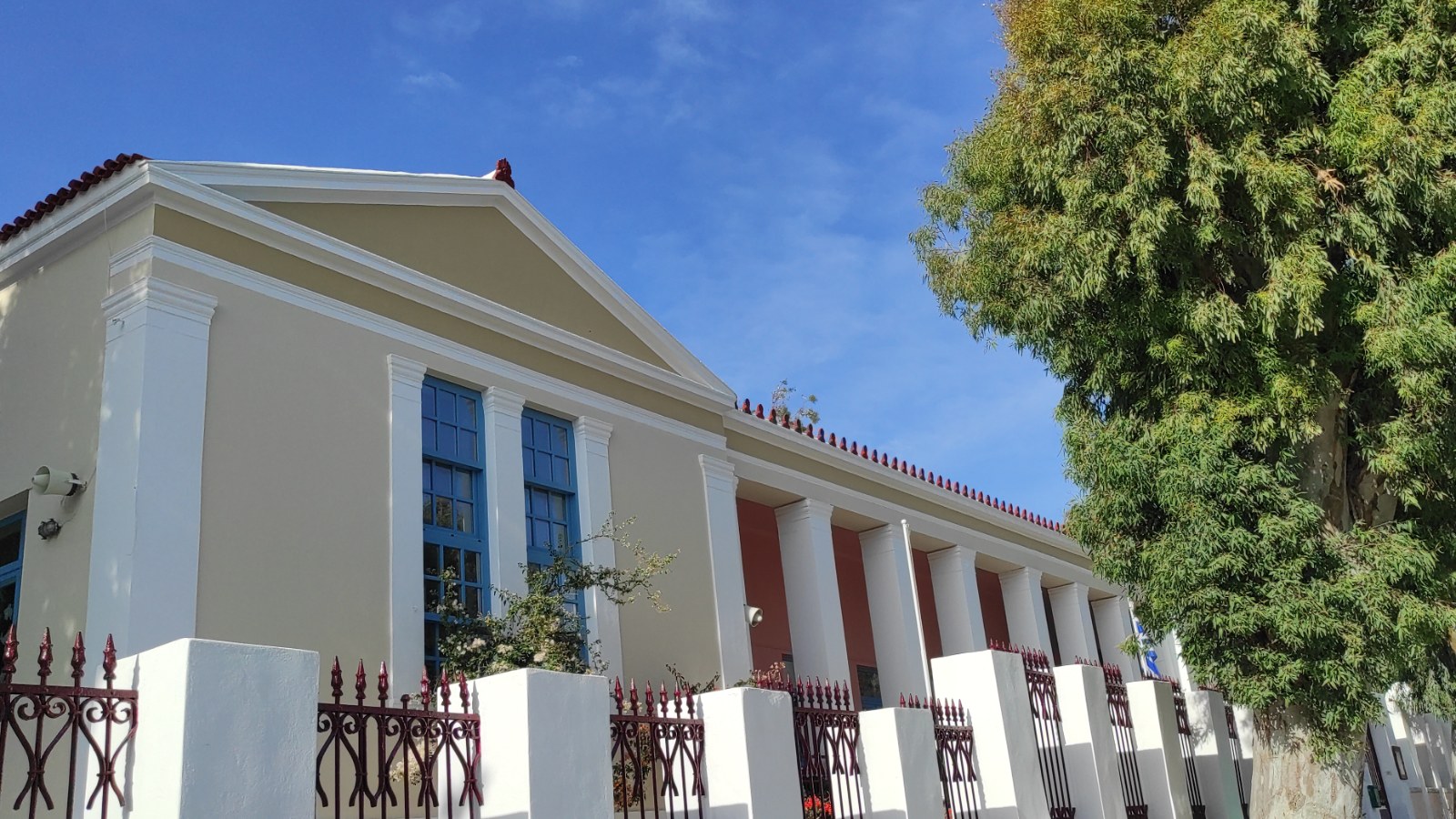 Σχολικό κτήριο στην Τήνο, νεοκλασικό με κολόνες και μπλε παράθυρα, πίσω από τα κάγκελα της αυλής