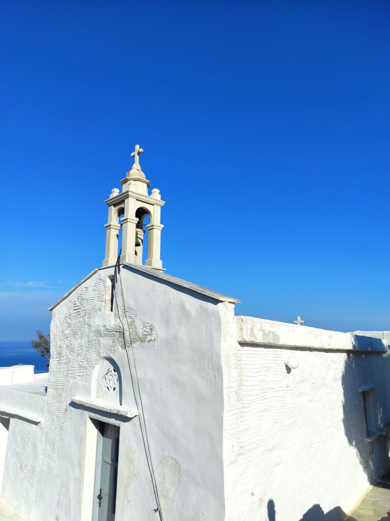 Λευκό εκκλησάκι με μαρμάρινο καμπαναριό κάτω από βαθύ γαλάζιο ουρανό, με θέα τη θάλασσα της Τήνου